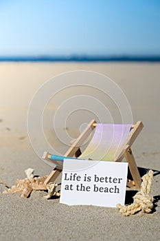 LIFE IS BETTER AT THE BEACH text on paper greeting card on background of beach chair lounge starfish summer vacation