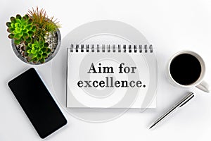 Life or business purpose concept - Aim for excellence. Text on a book with phone, cup of coffee, pen and plant on white background