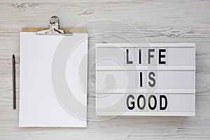 `Life is good` words on a lightbox, clipboard with blank sheet of paper on a white wooden surface, top view. Overhead, from abov