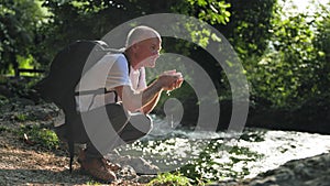 life in retirement, an elderly man with a backpack enjoys a walk and washes his face in river while sitting on bank in