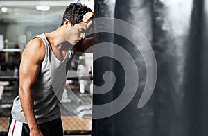 Life always throws him a punch. A muscular young man leaning against a boxing bag and looking defeated.