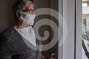 The life under Italy`s coronavirus lockdown. Senior woman with mask looking worriedly out of the window. Brescia Lombardy