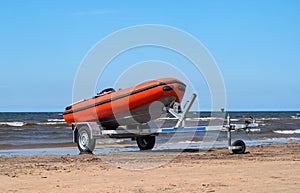 Lifeboat on trailer. The lifeboat is orange. The trailer with the boat is on the shore by the water. Sandy beach