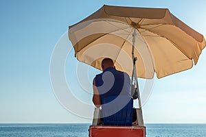 A lifeguard on the beach sits in a chair under a sun umbrella
