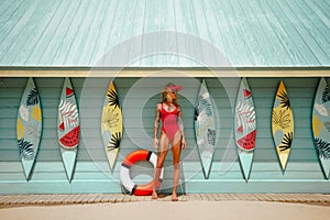 Lifeguard woman in red bikini and visor with lifebuoy on the sandy beach against surf station. Surfer female