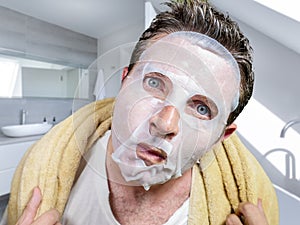 Young scared and surprised man at home using beauty paper facial mask cleansing doing anti aging facial treatment looking himself