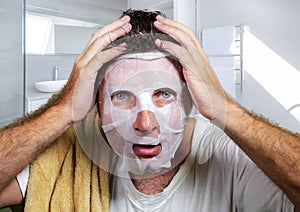 Young scared and surprised man at home using beauty paper facial mask cleansing doing anti aging facial treatment looking himself