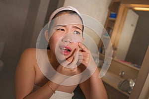 Lifestyle face portrait of young upset and expressive Asian Japanese woman squeezing pimples while looking at the mirror in the