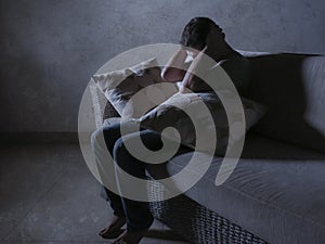 Lifestyle dramatic light portrait of young sad and depressed man sitting at shady home couch in pain and depression feeling stress