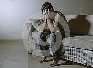 Lifestyle dramatic light portrait of young sad and depressed man sitting at shady home couch in pain and depression feeling stress