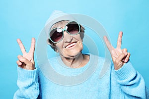 Lifestyle, emotion and people concept: Funny old lady wearing blue sweater, hat and sunglasses showing victory sign.