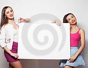 Lifestyle, emotion and people concept: Two young girl friends standing together and holding a blank billboard