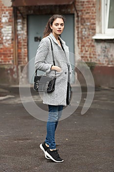 Lifestyle fashion portrait of beautiful young brunette woman in grey coat with black leather bag posing on street cloudy day