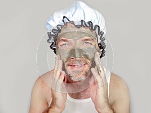 Lifestyle funny portrait of happy weird man on shower cap looking to himself in bathroom mirror with green cream on his face apply