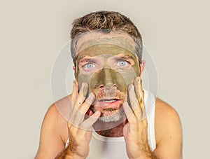 Attractive and surprised funny man horrified looking himself on bathroom mirror ugly and weird applying green cream on his face i