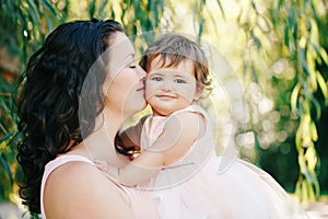 Lifestyle group portrait of beautiful white Caucasian brunette mother holding hugging daughter in pink dress kissing her in cheek