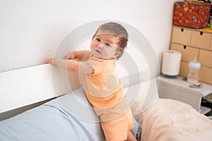 lifestyle home portrait of adorable and beautiful 9 months old baby girl learning to stand up by holding bed frame smiling