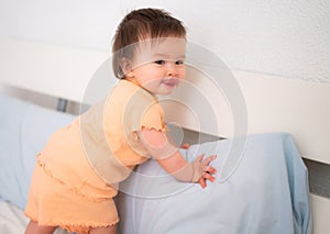 lifestyle home portrait of adorable and beautiful 9 months old baby girl learning to stand up by holding bed frame smiling