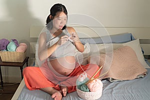 Lifestyle home portrait of young happy and beautiful Asian Chinese woman knitting clothes for the new baby relaxed in her bedroom