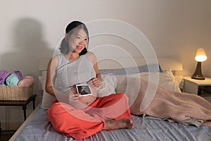 Lifestyle home portrait of young happy and beautiful Asian Chinese woman pregnant sitting on bed holding ultrasound photo excited