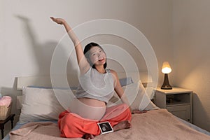 Lifestyle home portrait of young happy and beautiful Asian Chinese woman pregnant sitting on bed holding ultrasound photo excited