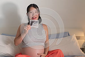 Lifestyle home portrait of young happy and beautiful Asian Chinese woman pregnant sitting on bed holding ultrasound photo excited