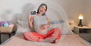 Lifestyle home portrait of young happy and beautiful Asian Chinese woman pregnant sitting on bed holding ultrasound photo excited