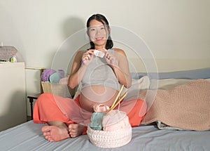 Lifestyle home portrait of young happy and beautiful Asian Japanese woman knitting clothes for the new baby relaxed in her bedroom