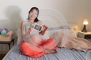 Lifestyle home portrait of young happy and beautiful Asian Japanese woman pregnant sitting on bed holding ultrasound photo excited