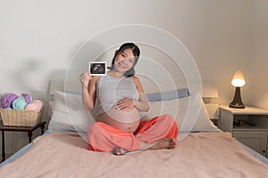 Lifestyle home portrait of young happy and beautiful Asian Japanese woman pregnant sitting on bed holding ultrasound photo excited