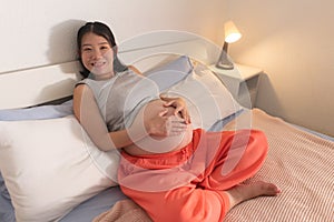 Lifestyle home portrait of young happy and beautiful Asian Korean woman pregnant sitting on bed relaxed and excited about