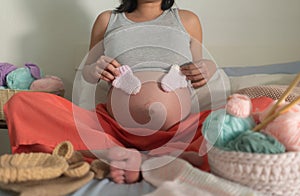 Lifestyle home portrait of young happy pregnant woman holding baby boots relaxed in her bedroom in maternity and handmade knitting