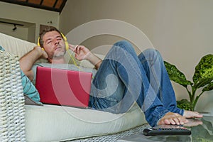 Lifestyle indoors portrait of young happy relaxed and attractive man lying at home sofa couch with headset and laptop listening to