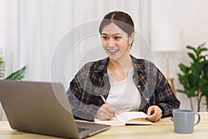 Lifestyle in living room concept, Young Asian woman using laptop and taking notes data on notebook