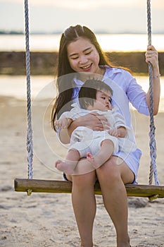 Young sweet and happy Asian Chinese woman holding baby girl swinging together at beach swing on Summer sunset in mother and little