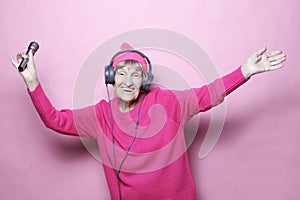 Lifestyle and people concept: Funny old lady listening music with headphones and singing with mic over pink background