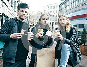 Lifestyle and people concept: two girls and guy eating fast food on city street together having fun, drinking coffee