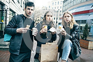 Lifestyle and people concept: two girls and guy eating fast food on city street together having fun, drinking coffee