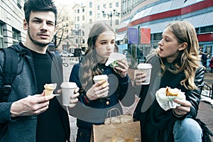 Lifestyle and people concept: two girls and guy eating fast food on city street together having fun, drinking coffee