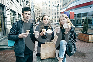 Lifestyle and people concept: two girls and guy eating fast food on city street together having fun, drinking coffee