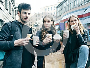 Lifestyle and people concept: two girls and guy eating fast food on city street together having fun, drinking coffee