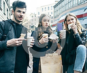 Lifestyle and people concept: two girls and guy eating fast food on city street together having fun, drinking coffee