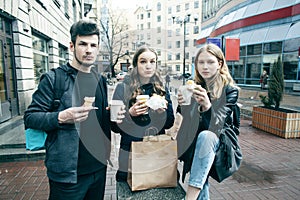 Lifestyle and people concept: two girls and guy eating fast food on city street together having fun, drinking coffee