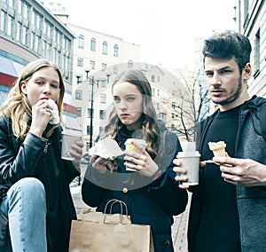 Lifestyle and people concept: two girls and guy eating fast food on city street together having fun, drinking coffee