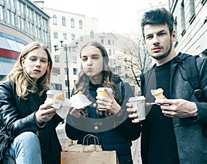 Lifestyle and people concept: two girls and guy eating fast food on city street together having fun, drinking coffee