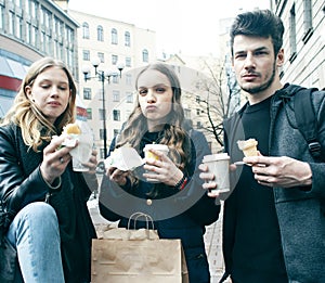 Lifestyle and people concept: two girls and guy eating fast food on city street together having fun, drinking coffee