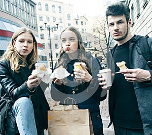 Lifestyle and people concept: two girls and guy eating fast food on city street together having fun, drinking coffee