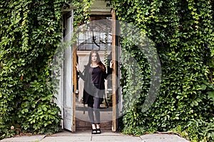 Lifestyle portrait of girl wearing blank black t-shirt, jeans and coat posing against building covered with green leaves