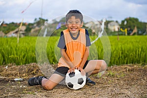 Lifestyle portrait of handsome and happy young boy holding soccer ball playing football outdoors at green grass field smiling chee