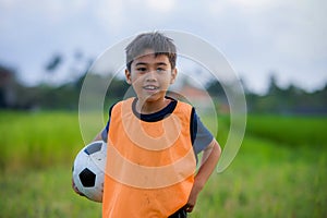 Lifestyle portrait of handsome and happy young boy holding soccer ball playing football outdoors at green grass field smiling chee